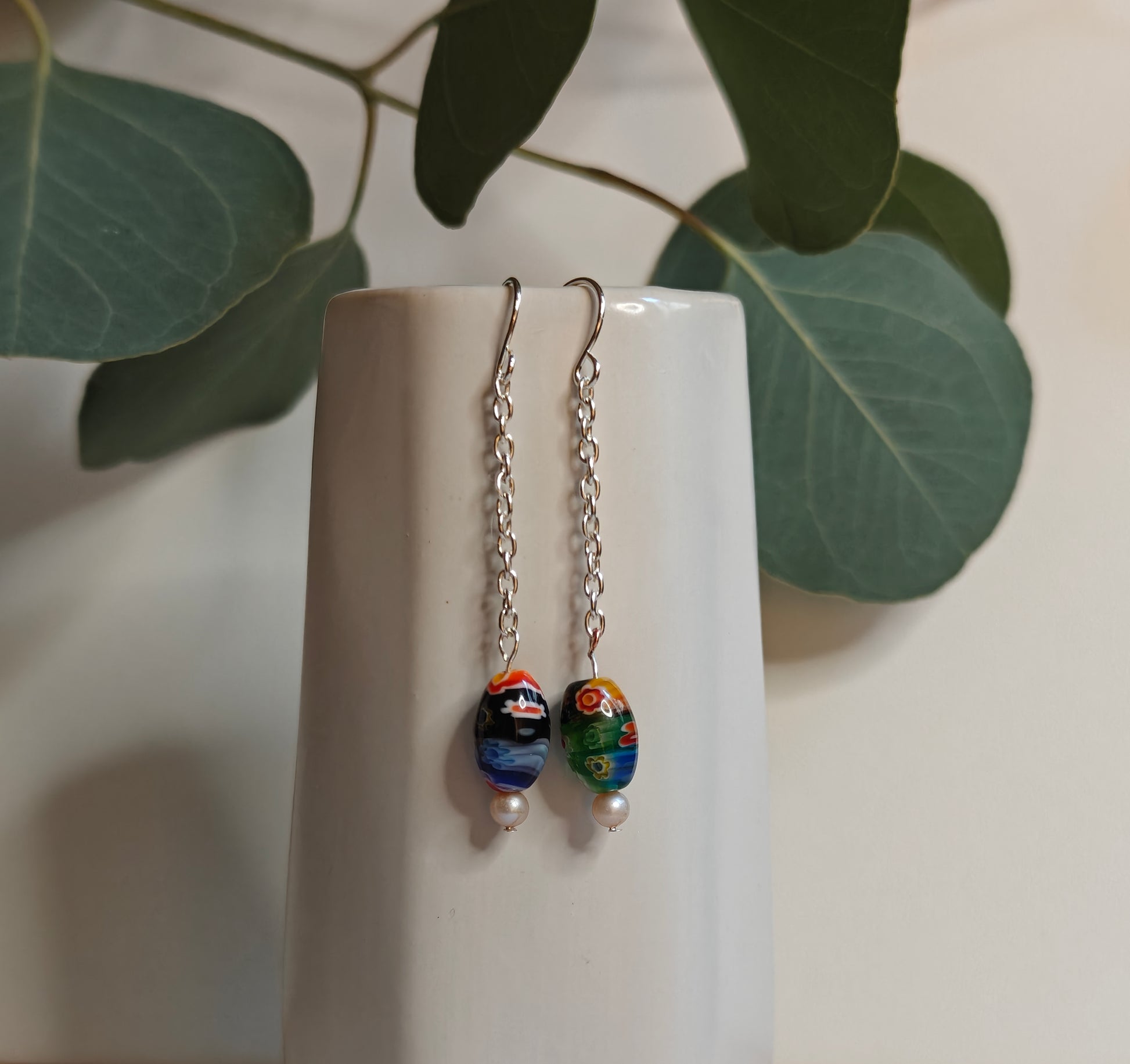 Image of the Silver toned Murano glass Earrings hanging off the side of a small white ceramic vase. It features a single colorful oval Murano glass bead, a small faux pearl bead and silver color earrings components 