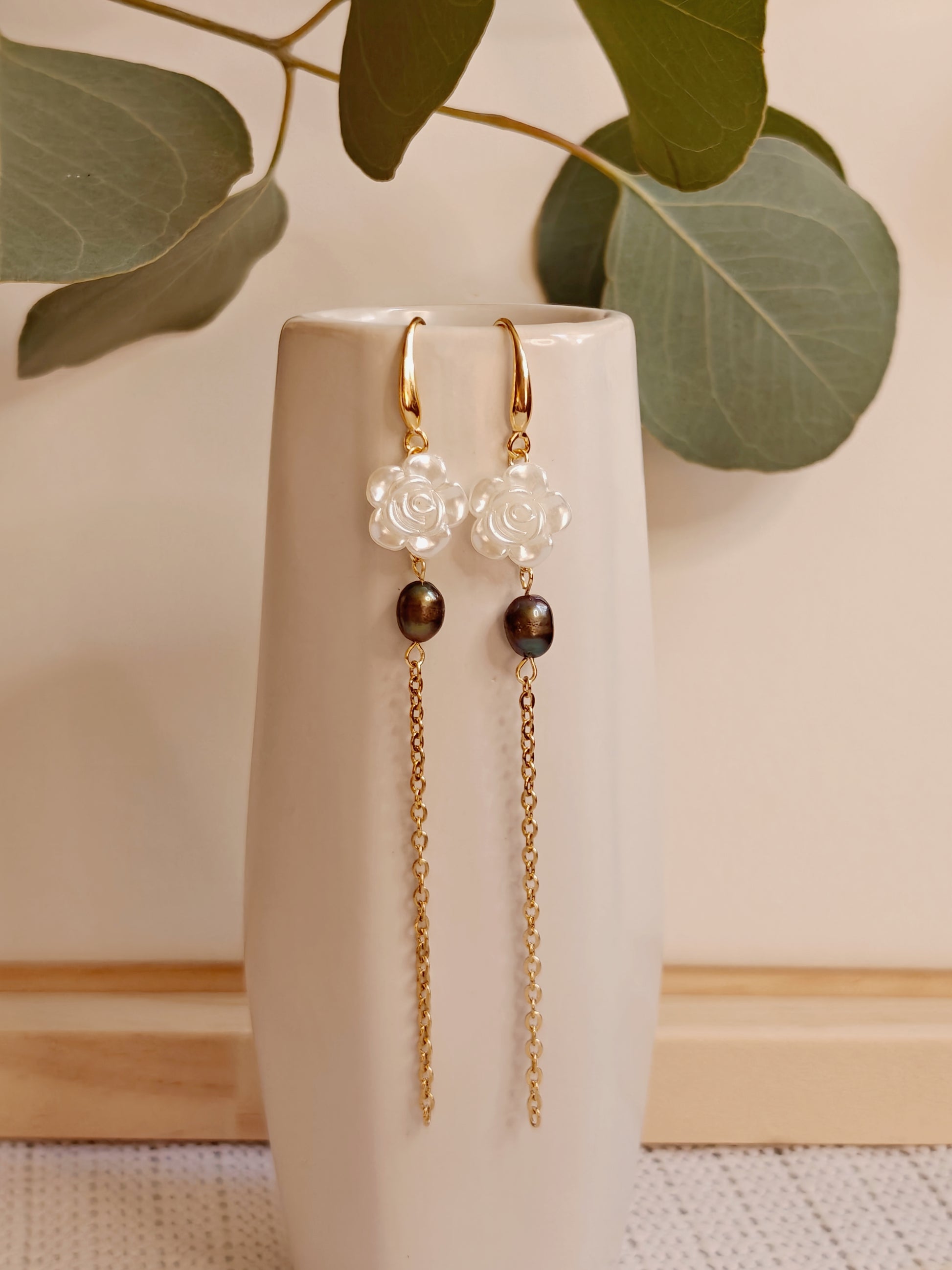 Image of the earrings hanging on the side of a small white ceramic vase. It is gold toned, featuring a white acrylic rose charm and a dark cultured fresh water pearl and finally a long gold color cable chain is attached to the bottom of the charms