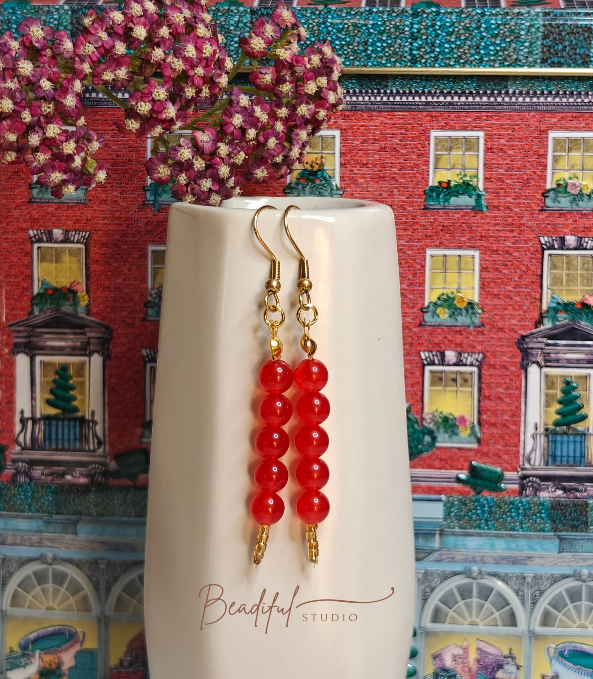 Image of the Tanghulu Earrings hanging off the side of a small white ceramic vase. It features five red beads and gold color earrings components 