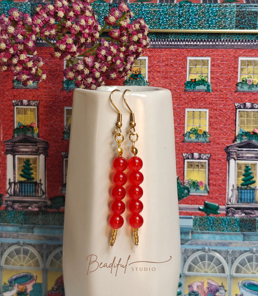 Image of the Tanghulu Earrings hanging off the side of a small white ceramic vase. It features five red beads and gold color earrings components 