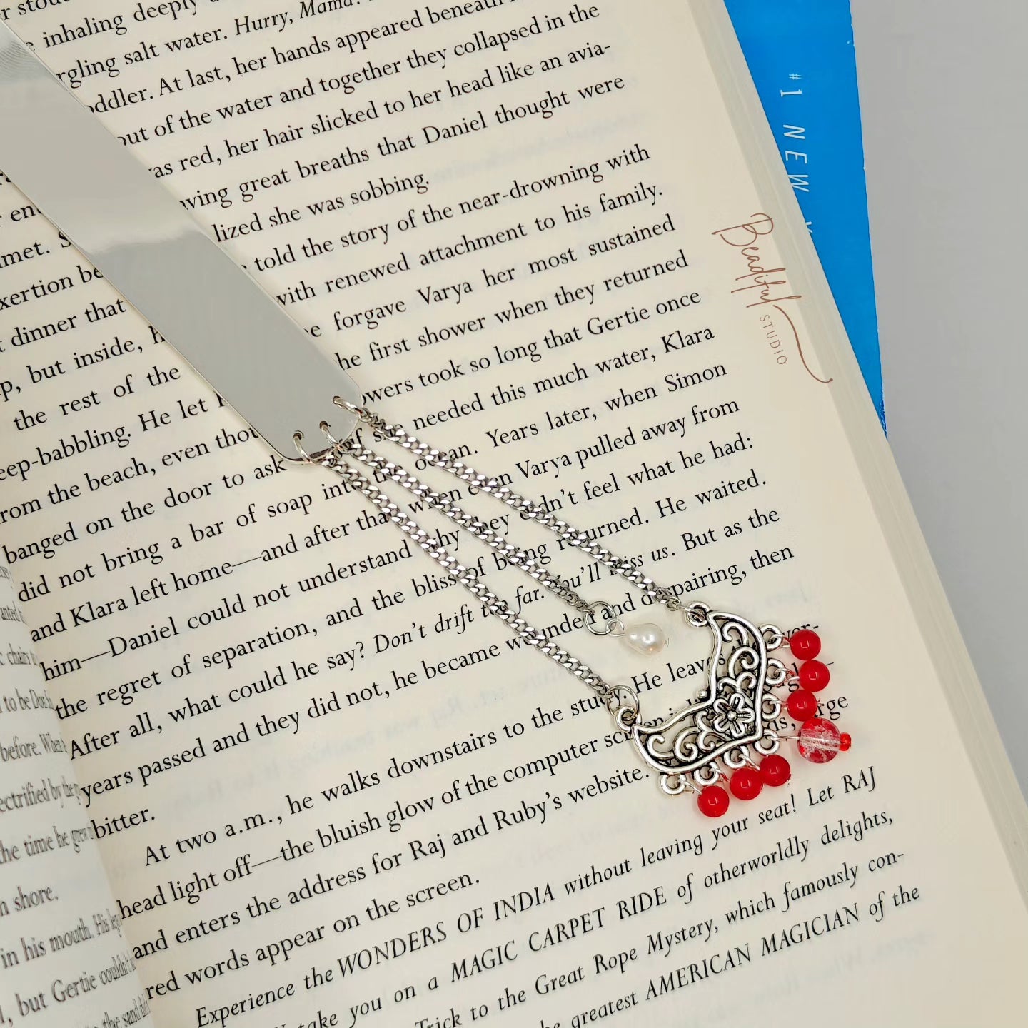 Image of the Lady Elara bookmark that is silver toned, laying flat on a page of a book. It features a white cultured freshwater pearl, and a bigger connector that attaches seven red color beads.