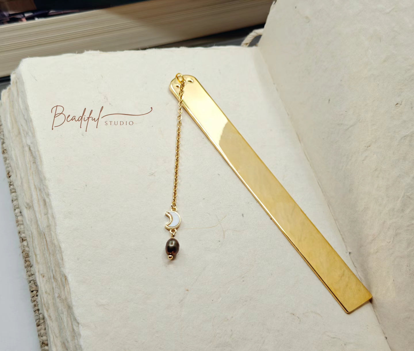 Image of the Luna Bookmark which features a gold color bar with a half moon charm and dark cultured freshwater pearl attached to it using a long cable chain.