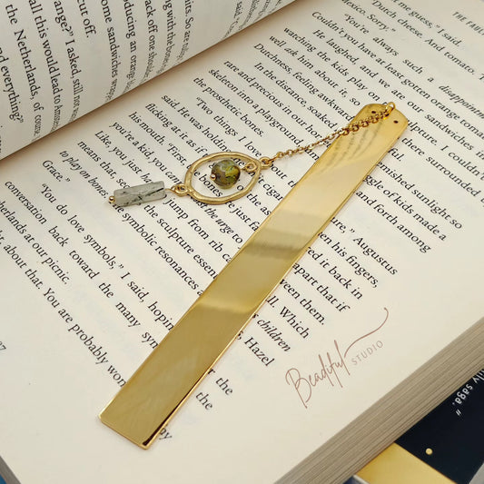 Image of a gold plated stainless steel bookmark laying flat on a book. It is attached with a two-inch gold plated cable chain and a Chinese inspired charm design that dangles from the bookmark.